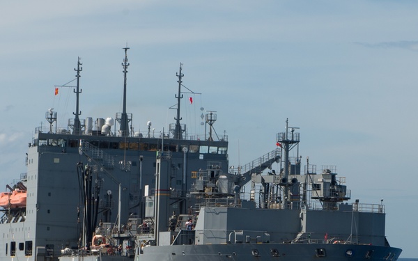 A large gray military auxiliary or logistics support vessel is underway on open blue ocean waters. The ship appears to be conducting vertical replenishment (VERTREP) operations, as evidenced by a helicopter visible in the background carrying a suspended load. The vessel is equipped with multiple masts, communication arrays, and deck equipment consistent with a naval supply or combat stores ship.
