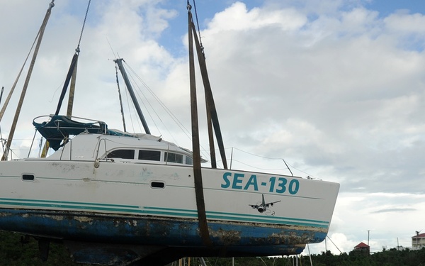 A catamaran labeled 'SEA-130' is being lifted by crane straps above a harbor filled with damaged and partially sunken sailboats, suggesting post-storm or post-hurricane salvage operations. Multiple vessels are visibly wrecked, capsized, or partially submerged in the calm harbor waters. The scene indicates significant marine disaster recovery efforts in a tropical or subtropical location.