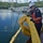 Two personnel, likely U.S. Coast Guard members based on their uniforms and American flag patch, are conducting what appears to be an oil spill response operation at a marina. They are handling yellow oil containment boom/absorbent materials on a dock, with several sailboats visible in the background, some appearing damaged or listing. The setting appears to be a tropical marina, possibly in the U.S. Virgin Islands or a similar Caribbean location.