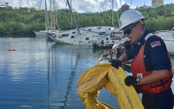 Two personnel, likely U.S. Coast Guard members based on their uniforms and American flag patch, are conducting what appears to be an oil spill response operation at a marina. They are handling yellow oil containment boom/absorbent materials on a dock, with several sailboats visible in the background, some appearing damaged or listing. The setting appears to be a tropical marina, possibly in the U.S. Virgin Islands or a similar Caribbean location.