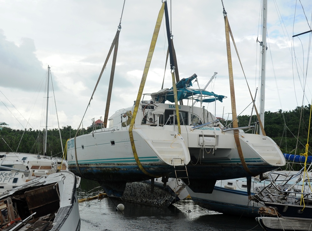 A damaged catamaran sailboat is being lifted by a crane using heavy yellow straps in what appears to be a post-hurricane marina recovery operation. The vessel, named 'SEA-[?]0' and hailing from Albuquerque, NM, shows significant damage including marine growth on the hull, suggesting it was submerged or grounded. Surrounding the lifted catamaran are multiple other damaged and partially sunken sailboats in a crowded marina.