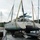 A damaged catamaran sailboat is being lifted by a crane using heavy yellow straps in what appears to be a post-hurricane marina recovery operation. The vessel, named 'SEA-[?]0' and hailing from Albuquerque, NM, shows significant damage including marine growth on the hull, suggesting it was submerged or grounded. Surrounding the lifted catamaran are multiple other damaged and partially sunken sailboats in a crowded marina.