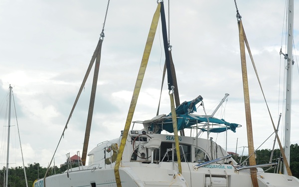 A damaged catamaran sailboat is being lifted by a crane using heavy yellow straps in what appears to be a post-hurricane marina recovery operation. The vessel, named 'SEA-[?]0' and hailing from Albuquerque, NM, shows significant damage including marine growth on the hull, suggesting it was submerged or grounded. Surrounding the lifted catamaran are multiple other damaged and partially sunken sailboats in a crowded marina.