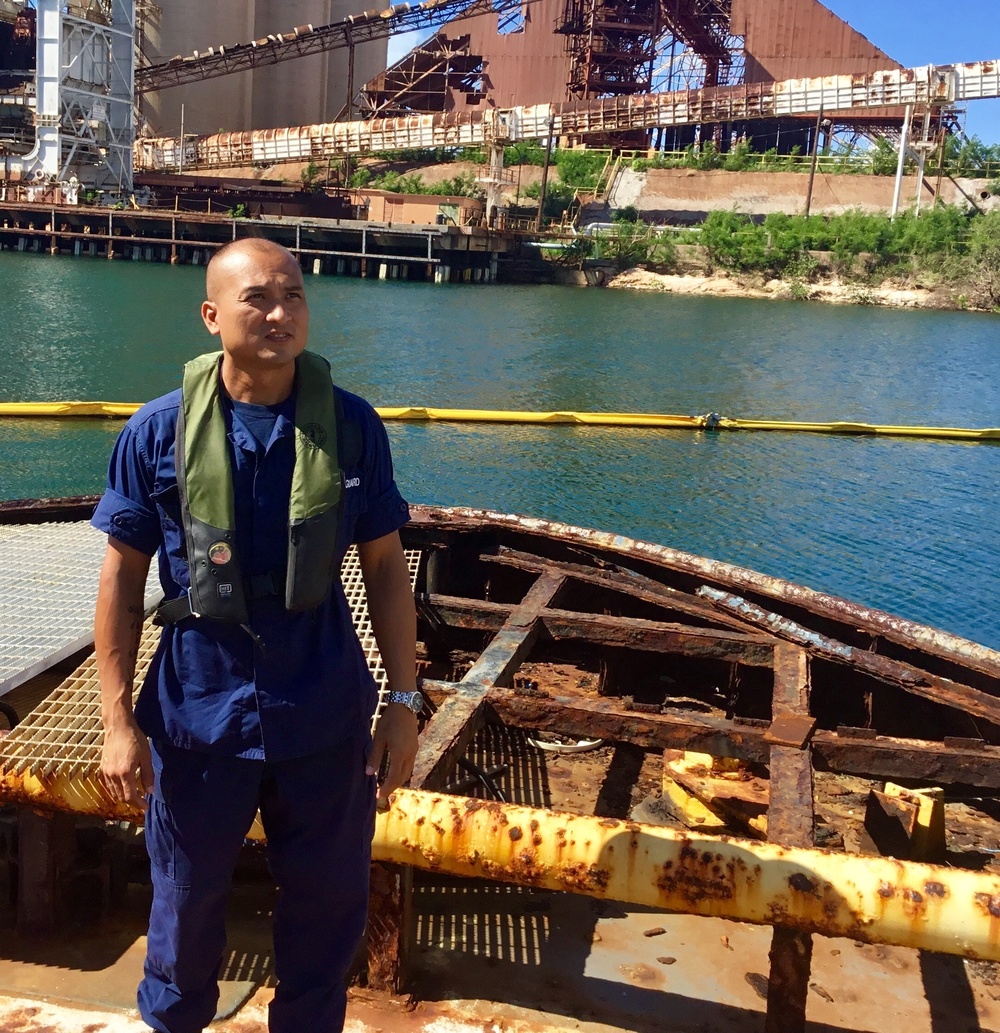 A man wearing blue coveralls and a green inflatable personal flotation device (PFD) stands on a heavily rusted and deteriorated vessel or barge structure near an industrial waterway. Behind him, a yellow oil containment boom is visible floating on the water, along with large industrial port infrastructure showing significant rust and age. The setting appears to be an inspection or assessment of a derelict vessel at an industrial port facility.