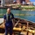 A man wearing blue coveralls and a green inflatable personal flotation device (PFD) stands on a heavily rusted and deteriorated vessel or barge structure near an industrial waterway. Behind him, a yellow oil containment boom is visible floating on the water, along with large industrial port infrastructure showing significant rust and age. The setting appears to be an inspection or assessment of a derelict vessel at an industrial port facility.