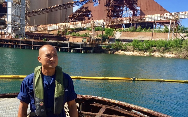 A man wearing blue coveralls and a green inflatable personal flotation device (PFD) stands on a heavily rusted and deteriorated vessel or barge structure near an industrial waterway. Behind him, a yellow oil containment boom is visible floating on the water, along with large industrial port infrastructure showing significant rust and age. The setting appears to be an inspection or assessment of a derelict vessel at an industrial port facility.
