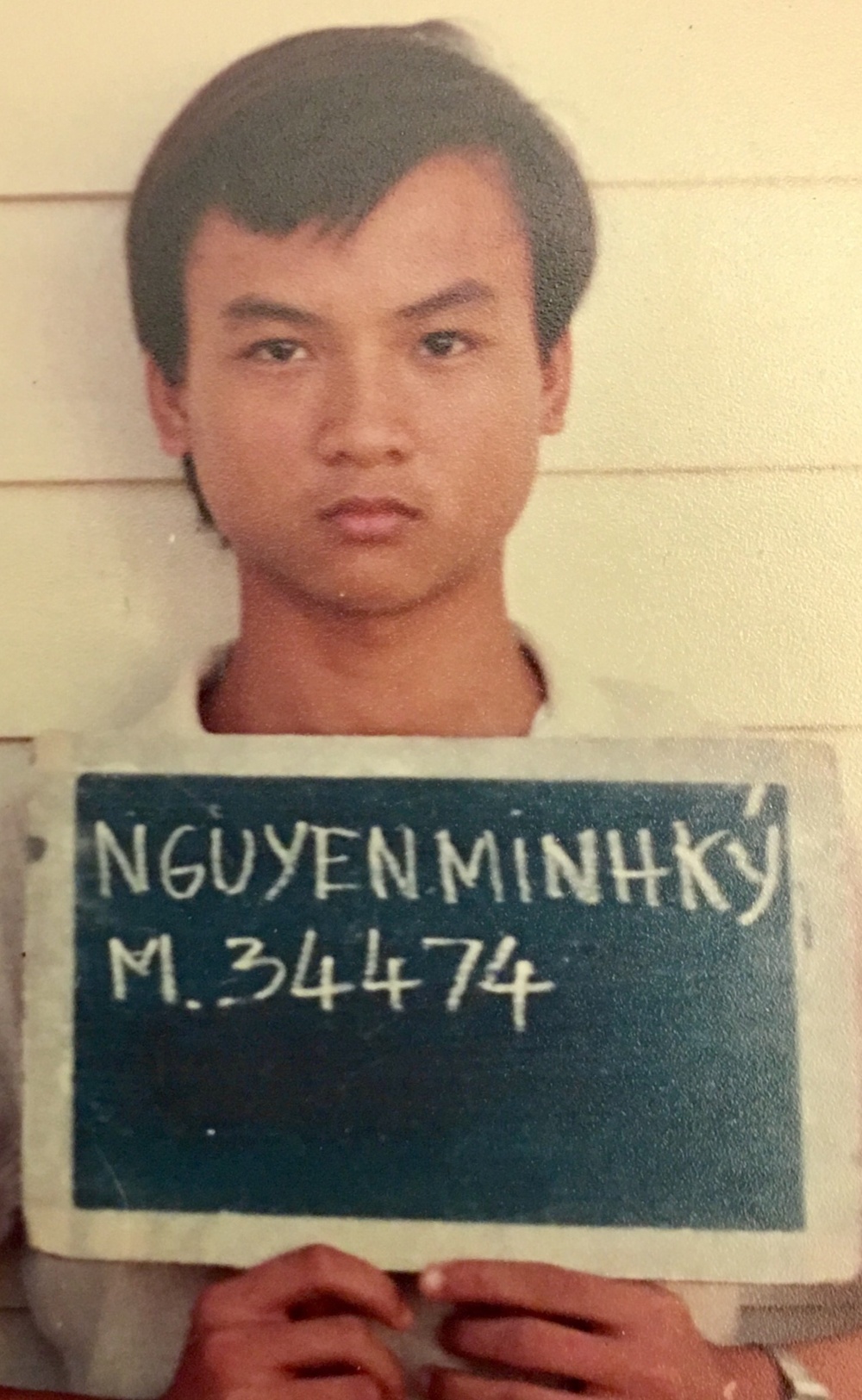 This is an archival identification photograph, consistent with a booking or registration photo. The subject is holding a dark board with handwritten text reading 'NGUYEN MINH KY' and 'M.34474'. The photo appears aged, suggesting it was taken several decades ago.
