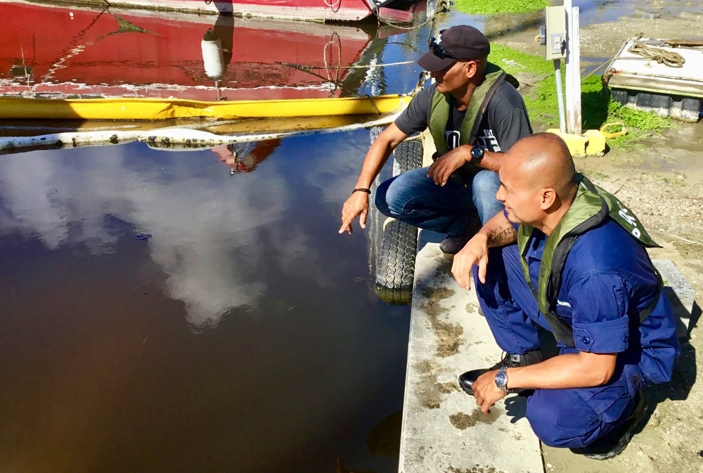 Two men wearing olive-green life vests are crouching at the edge of a dock or pier, examining what appears to be an oil spill or heavily contaminated dark water. One man in a black cap is pointing toward the water surface while the other, in a blue uniform, observes closely. The setting appears to be a small harbor or marina with red and yellow boats visible in the background.
