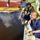 Two men wearing olive-green life vests are crouching at the edge of a dock or pier, examining what appears to be an oil spill or heavily contaminated dark water. One man in a black cap is pointing toward the water surface while the other, in a blue uniform, observes closely. The setting appears to be a small harbor or marina with red and yellow boats visible in the background.