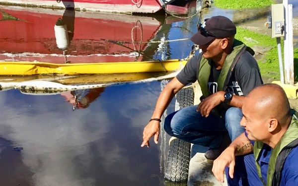 Two men wearing olive-green life vests are crouching at the edge of a dock or pier, examining what appears to be an oil spill or heavily contaminated dark water. One man in a black cap is pointing toward the water surface while the other, in a blue uniform, observes closely. The setting appears to be a small harbor or marina with red and yellow boats visible in the background.