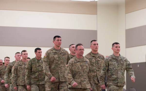 A formation of U.S. Army soldiers in Operational Camouflage Pattern (OCP) uniforms marches in an indoor facility, likely during a formal military ceremony or mobilization event. The soldiers are arranged in multiple columns and are moving in a coordinated manner across a polished floor. The setting appears to be a large gymnasium or event hall.