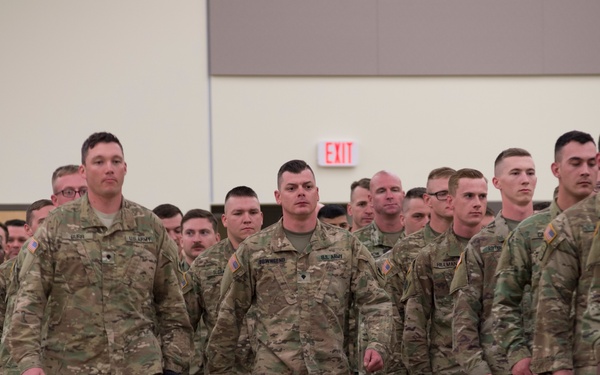 A large formation of U.S. Army soldiers in Operational Camouflage Pattern (OCP) uniforms marching in an indoor facility, likely a gymnasium or assembly hall. The soldiers appear to be participating in a formal military ceremony or formation event. American flag patches are visible on their shoulders, identifying them as U.S. Army personnel.
