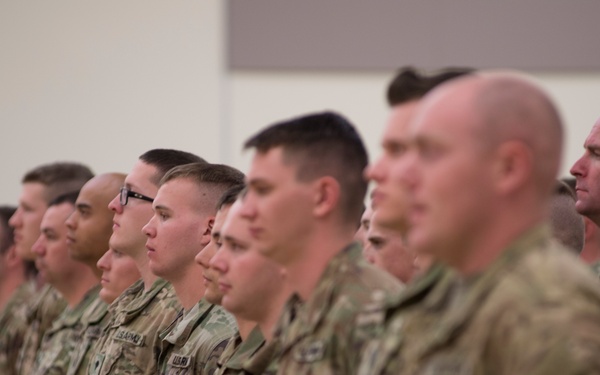 A large formation of military personnel standing at attention in what appears to be an indoor facility. All individuals are wearing U.S. Army Operational Camouflage Pattern (OCP) uniforms, suggesting this is a formal military ceremony or formation. The soldiers are arranged in multiple rows and appear to be facing the same direction.