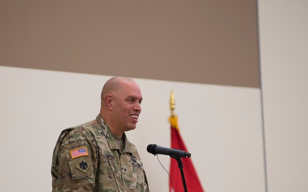 A U.S. Army officer in OCP (Operational Camouflage Pattern) uniform is speaking at a podium with a microphone during what appears to be a formal military ceremony. A red general officer flag with one white star is visible in the background, indicating the presence of a Brigadier General. A second soldier in uniform is seated in the background.