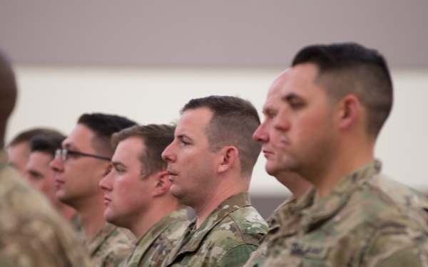 A group of military personnel in U.S. Army OCP (Operational Camouflage Pattern) uniforms are standing in formation, likely during a ceremony or official event held indoors. The soldiers are standing at attention or parade rest, facing the same direction in a disciplined line. A small civilian audience is partially visible in the background to the right.