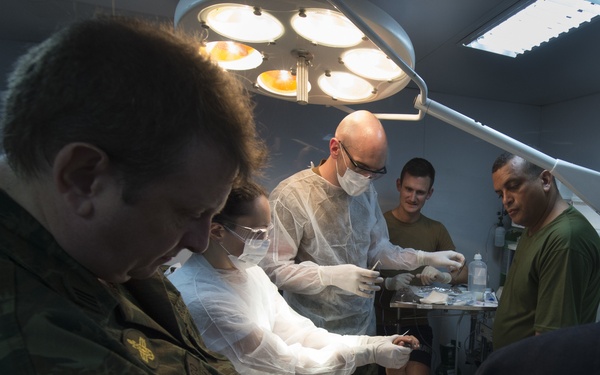 A medical procedure is being performed on a patient lying on an operating table under surgical lighting. Multiple personnel are present, including at least one individual wearing a Brazilian military uniform with a 'BRASIL' patch and 'Medicina Operativa' insignia. The setting appears to be a military medical facility or field surgical unit.