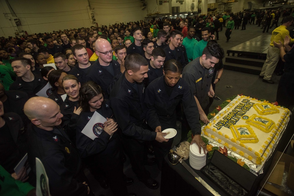 A large group of military personnel in dark uniforms are gathered in what appears to be a ship's hangar bay for a celebration ceremony. A large decorated cake with yellow frosting text reading 'Congratulations on Your Achievement' is being served at the front. The crowd extends deep into the background, suggesting a significant number of attendees at this recognition event.