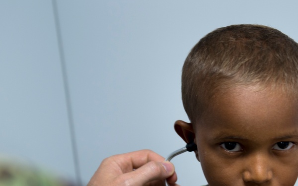 A military medical personnel in camouflage uniform is examining a young child using a stethoscope. The child, wearing a yellow-green shirt, looks directly at the camera with a serious expression. The setting appears to be a medical examination environment with a light blue/gray background.
