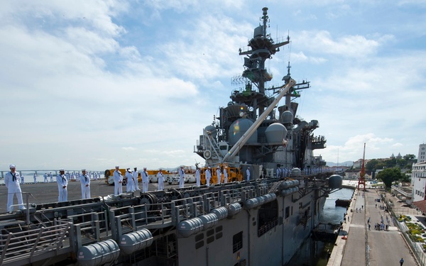 A large US Navy amphibious assault ship is docked at a port, with sailors in white dress uniforms manning the rails along the flight deck. The vessel is berthed alongside a civilian pier in what appears to be an urban port environment, with city buildings visible in the background. The scene suggests a formal port arrival or departure ceremony.
