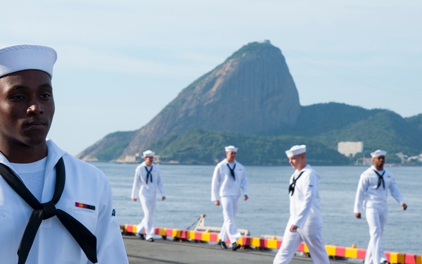 A group of U.S. Navy sailors in dress white uniforms are stationed on the deck of a naval vessel, with the iconic Sugarloaf Mountain (Pão de Açúcar) of Rio de Janeiro, Brazil visible in the background. The foreground features a young sailor standing at attention, while several others walk along the ship's deck behind him. The scene appears to depict a formal naval port visit or ceremonial manning of the rails.