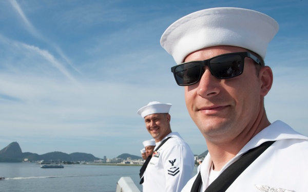 Three U.S. Navy sailors in dress white uniforms are standing on the deck of a naval vessel in what appears to be a harbor, with a distinctive mountain landmark visible in the background consistent with Rio de Janeiro, Brazil. The foreground sailor displays multiple service ribbons, a warfare qualification badge, aviator wings, and a Petty Officer First Class rating badge on his sleeve. The image is taken in clear daylight with calm water and blue skies.