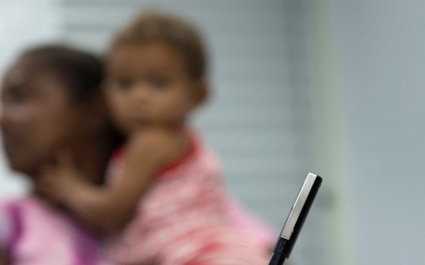 A person in military camouflage uniform is writing on a document or form in the foreground. In the blurred background, an adult woman in a pink shirt is holding a young child. The scene appears to depict a military humanitarian assistance or administrative processing event.