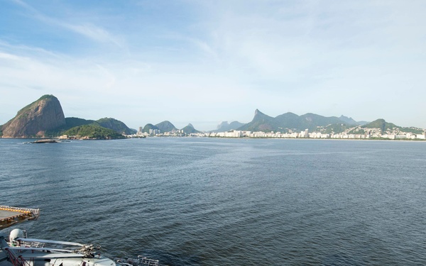 The image shows the flight deck of a US Navy vessel anchored in what appears to be Guanabara Bay, Rio de Janeiro, Brazil, with the iconic Sugarloaf Mountain (Pão de Açúcar) and Christ the Redeemer statue visible in the background. At least two military helicopters are parked on the deck, and a sailor in white dress uniform is visible walking on the starboard side. The word 'NAVY' is partially visible on the right side of the frame.