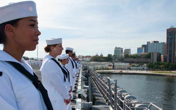 A line of U.S. Navy sailors in white dress uniforms stand at attention along the edge of the flight deck of USS Wasp (LHD-1) during a port visit to what appears to be Rio de Janeiro, Brazil. The sailors are performing a 'manning the rails' ceremonial formation, a traditional naval custom during port arrivals or departures. The city skyline, waterfront buildings, and a harbor crane are visible in the background across the water.