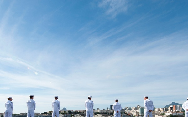 A line of naval personnel dressed in white dress uniforms stand at attention along the edge of what appears to be an aircraft carrier flight deck. They are facing outward toward a harbor cityscape with buildings, mountains, and other vessels visible in the background. The formation suggests a ceremonial 'manning the rails' procedure, typically performed during port arrivals or departures.