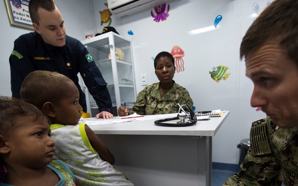 A military medical outreach scene showing U.S. Navy personnel and a Brazilian military member providing medical care to young children in a pediatric-themed clinic room. A U.S. Navy service member in camouflage uniform is crouching down to interact with small children, while a female Navy personnel sits at a desk with a stethoscope, and a Brazilian military member stands nearby. The room is decorated with colorful sea creature wall decals, suggesting a child-friendly medical facility.