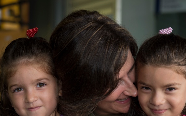 A woman with long dark hair embraces two young girls who appear to be twins or close-in-age sisters. The woman is leaning her head affectionately against the girl on the right, while holding both children close. The scene conveys warmth and familial affection.