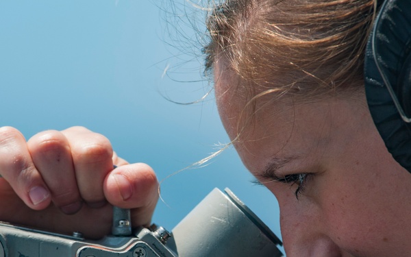 A person wearing military-style communication headphones is looking through a optical sighting or navigation instrument marked with the number '8'. The equipment appears to be mounted on a naval vessel, consistent with a shipboard alidade or bearing circle used for navigation. The image is taken outdoors under clear blue sky conditions.