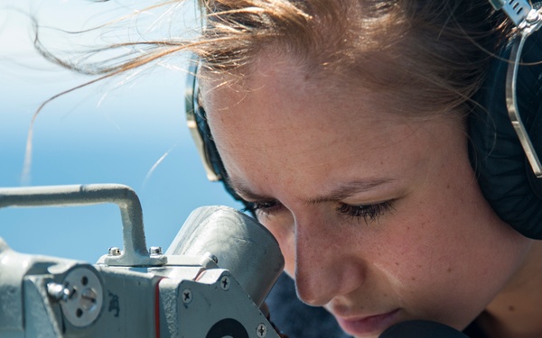 A young woman wearing military-style communication headphones is looking through a naval optical sighting or bearing instrument, marked with the number '8'. The setting appears to be an outdoor deck of a naval vessel, indicated by the equipment style and open sky background. Her hair is blowing in the wind, suggesting an open-air maritime environment.