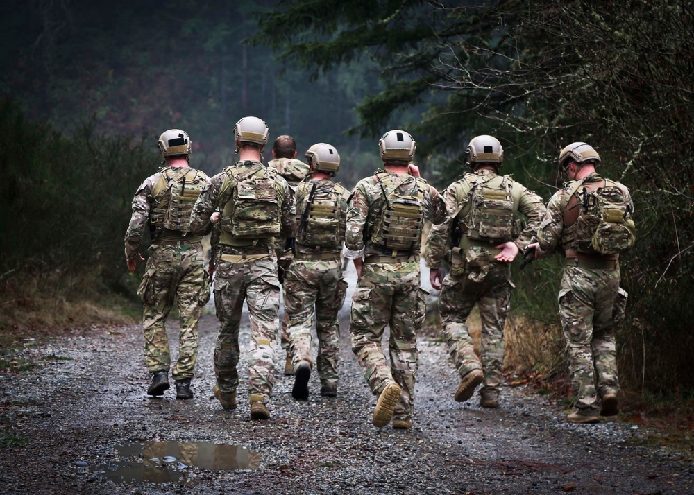 A group of approximately seven military personnel in full tactical gear are walking away from the camera along a gravel forest path. All individuals are wearing matching multicam camouflage uniforms, tan/khaki helmets, plate carriers, and tactical backpacks. The setting appears to be a wooded training or operational environment under overcast conditions.