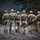 A group of approximately seven military personnel in full tactical gear are walking away from the camera along a gravel forest path. All individuals are wearing matching multicam camouflage uniforms, tan/khaki helmets, plate carriers, and tactical backpacks. The setting appears to be a wooded training or operational environment under overcast conditions.