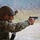 A military or law enforcement personnel is firing a semi-automatic pistol at what appears to be an outdoor shooting range. A spent brass casing is visibly ejecting from the firearm's ejection port, captured mid-air. The shooter is wearing full tactical gear including a combat helmet, camouflage uniform, eye protection, and hearing protection.