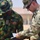 Two military personnel are reviewing a document together outdoors. One soldier wears a Nigerian Army uniform with the name tag 'A. OBOLO' and three-star insignia, while the other wears a MultiCam/OCP uniform with a SETAF (Southern European Task Force) shoulder patch. The interaction appears to be a formal or administrative exchange between allied military forces.