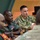 A meeting or briefing is taking place between U.S. Army personnel and foreign military officers seated around a table. The U.S. Army soldier in OCP (Operational Camouflage Pattern) uniform appears to be speaking or presenting, while officers in green dress uniforms with medal ribbons listen attentively. This appears to be a multinational military engagement or partnership meeting.