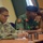 Three military personnel are seated at a conference table, appearing to be attending a meeting or briefing. The group includes a U.S. Army soldier in MultiCam/OCP uniform and two individuals in Nigerian Army dress uniforms. The setting suggests a multinational military cooperation or liaison event.