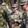 The image shows a group of military personnel in an outdoor setting engaged in what appears to be a briefing or coordination meeting. At least two U.S. Army soldiers in OCP (Operational Camouflage Pattern) uniforms are interacting with personnel wearing a different camouflage pattern, likely from a partner nation's military. One U.S. Army soldier, identified by the name tape 'MUNOZ' and captain rank insignia, is holding a notepad and appears to be taking notes or reviewing documents.