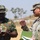 Three military personnel are engaged in a discussion outdoors, with two U.S. Army soldiers and one Nigerian Army soldier reviewing documents and taking notes. The interaction appears to be a formal briefing or coordination meeting, likely during a joint military exercise or training event. The setting is an open outdoor area with sparse vegetation visible in the background.