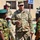 A U.S. Army soldier, identified by name tape as 'MONTEMAYOR' and wearing OCP (Operational Camouflage Pattern) uniform, is engaged in a briefing or discussion with several military personnel wearing Nigerian Army camouflage uniforms. The setting appears to be a military installation, with a sign reading 'BN HQ' (Battalion Headquarters) visible in the background. This appears to be a multinational military engagement or training exercise.