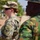 Two military personnel are standing outdoors in what appears to be a warm, arid environment. The foreground shows a female soldier in US Army MultiCam (OCP) uniform holding a spiral notebook, while a second soldier in green camouflage with a red and green flag patch stands to her right. The setting suggests a joint military exercise or cooperative engagement between two different national armed forces.