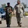 Two military personnel are walking together on a paved road, engaged in conversation. One wears a Nigerian Army uniform with a dark blue beret, and the other wears a US Army MultiCam (OCP) uniform with a patrol cap. The setting appears to be a military installation or base, with vehicles and trees visible in the background.