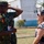 A Nigerian Army soldier in camouflage uniform is engaged in a friendly conversation with a civilian woman outdoors. The soldier is smiling and gesturing while the woman, wearing sunglasses and a black-and-white patterned top, appears to be laughing or reacting positively. The setting appears to be a military or government compound with white buildings and trees visible in the background.