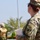 A U.S. Army soldier named Munoz, wearing Operational Camouflage Pattern (OCP) uniform, is photographed from behind/side holding a spiral notebook with handwritten notes. She is wearing sunglasses and a patrol cap, with a unit patch visible on her left sleeve. A second individual in a different military uniform is visible in the blurred background.