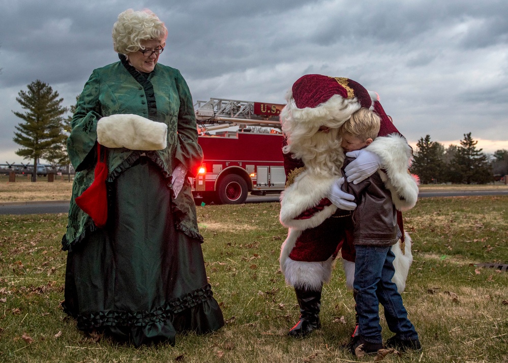 A Christmas-themed outdoor event featuring performers dressed as Santa Claus and Mrs. Claus interacting with a young child. Santa is bending down to embrace a small boy while Mrs. Claus, dressed in a green Victorian-style gown, watches warmly. A red fire truck with partial 'U.S.' lettering is visible in the background, suggesting this may be a military or government-sponsored holiday event.