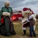 A Christmas-themed outdoor event featuring performers dressed as Santa Claus and Mrs. Claus interacting with a young child. Santa is bending down to embrace a small boy while Mrs. Claus, dressed in a green Victorian-style gown, watches warmly. A red fire truck with partial 'U.S.' lettering is visible in the background, suggesting this may be a military or government-sponsored holiday event.