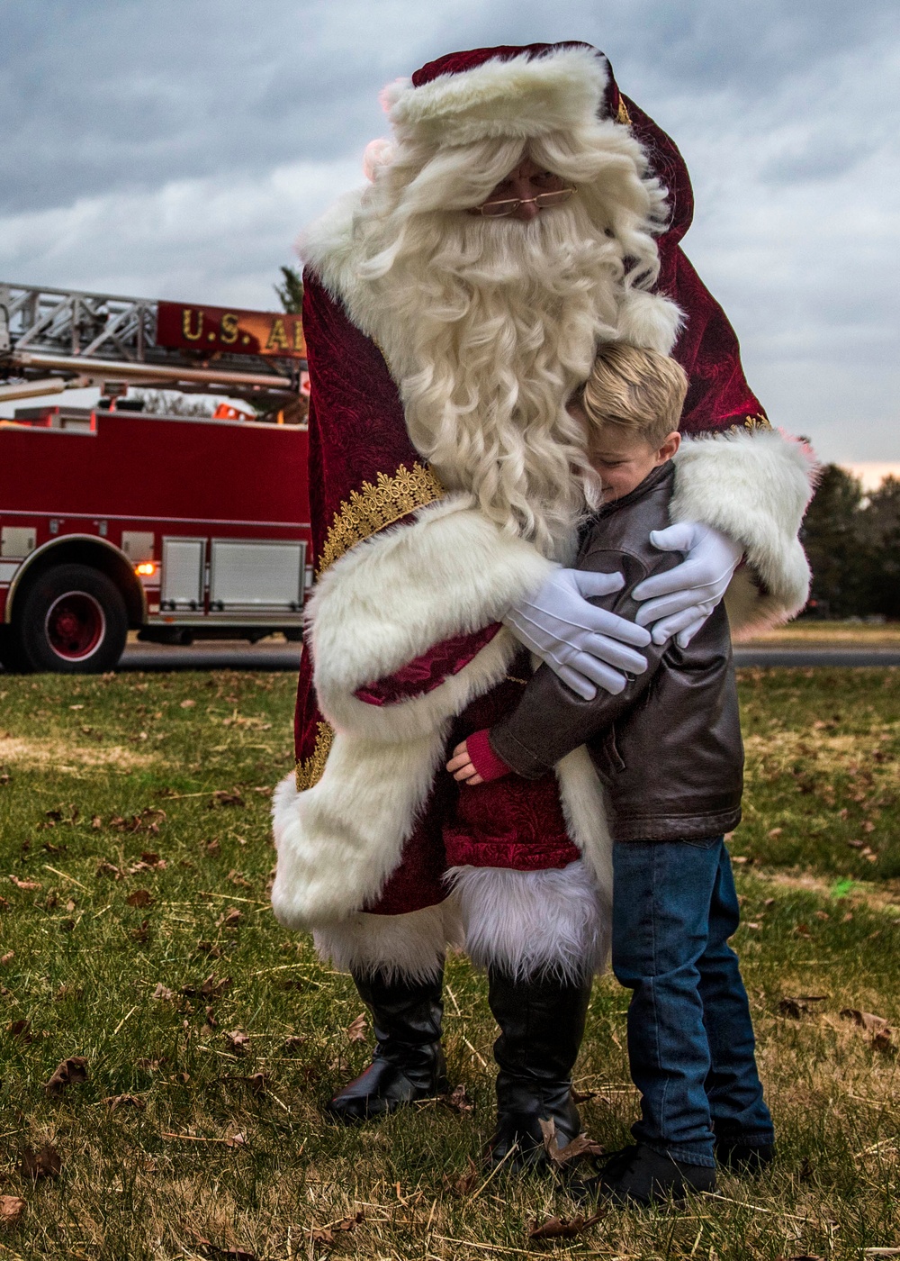 A person dressed as Santa Claus in a detailed dark red and white fur-trimmed costume is hugging a young boy outdoors on a grassy area. In the background, a red fire truck with partial text reading 'U.S. A[ir Force]' is visible, suggesting this is a military base holiday event. The scene appears to be a Christmas community event photographed under overcast skies.