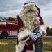 A person dressed as Santa Claus in a detailed dark red and white fur-trimmed costume is hugging a young boy outdoors on a grassy area. In the background, a red fire truck with partial text reading 'U.S. A[ir Force]' is visible, suggesting this is a military base holiday event. The scene appears to be a Christmas community event photographed under overcast skies.
